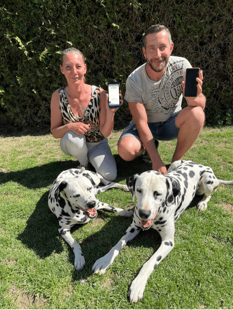 Die Pfotenportal Gruender Yvonne und Manuel zeigen im Garten mit ihren Dalmatinern den digitalen Gratis Hundepass auf einem Smartphone um die Notwendigkeit der Vorsorge zu betonen