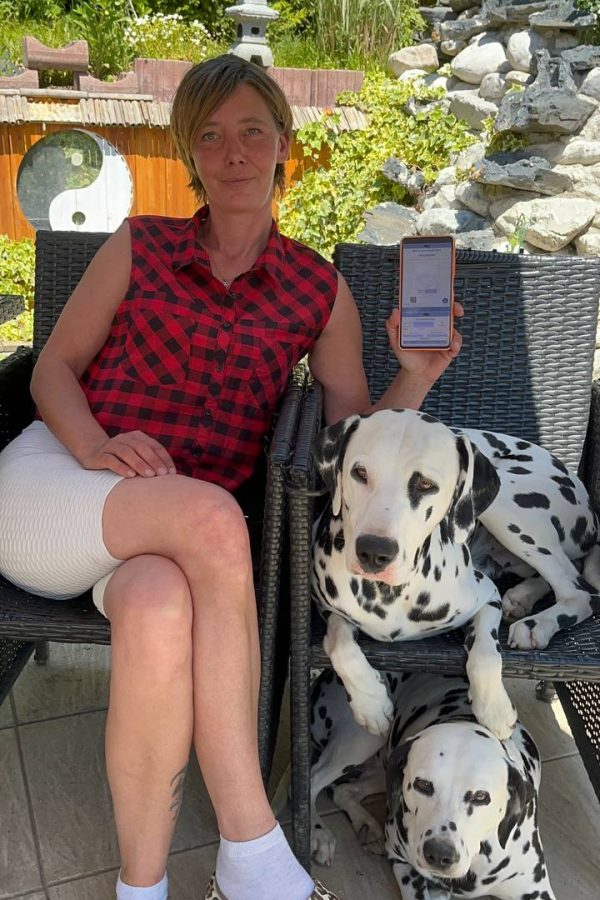 Yvonne vom Pfotenportal sitzt laechelnd im garten mit ihren Hunden Buddy und Jina und zeigt stolz den gratis digitalen Hundepass auf einem Smartphone-Bildschirm. Der Pass Hunde-Foto ist klar zu erkennen. Ein authentisches Vorschaubild für die kostenlose Sektion auf pfotenportal.at.